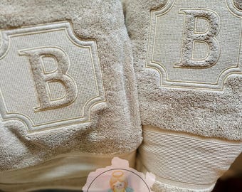 May include: Two beige bath towels with an embroidered letter "B" within a decorative frame. The towels have a plush texture and a striped border. The towels are part of the "Little Angel Embroidery" collection.