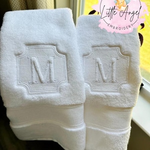 May include: Two white terry cloth towels featuring a centered embroidered monogram of the letter M within a decorative square frame. Each towel has a horizontal textured band near the bottom edge. A pink circular logo reads Little Angel Embroidery.