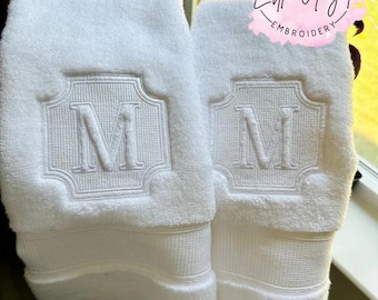 May include: Two white terry cloth towels featuring a centered embroidered monogram of the letter M within a decorative square frame. Each towel has a horizontal textured band near the bottom edge. A pink circular logo reads Little Angel Embroidery.