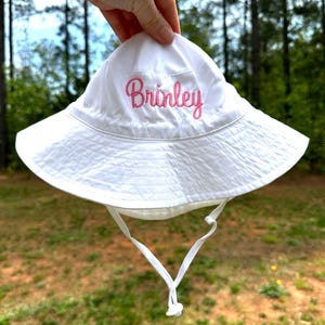 May include: White sun hat with a pink embroidered name "Brinley" on the front. The hat has a white drawstring.