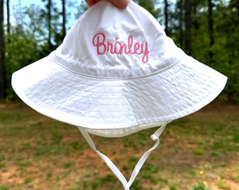 May include: White sun hat with a pink embroidered name "Brinley" on the front. The hat has a white drawstring.