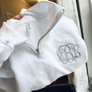 May include: White quarter-zip sweatshirt with a silver zipper and embroidered monogram. The sweatshirt has a classic design and is made of a soft material. The monogram is in a script font.