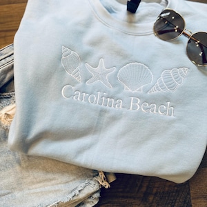 May include: Light blue sweatshirt with white embroidery of seashells and a starfish. The text "Carolina Beach" is embroidered below the shells. A pair of sunglasses is lying on top of the sweatshirt.