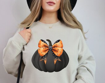 May include: Beige sweatshirt featuring a black pumpkin with an orange bow adorned with white stars. The bow has black polka dots. The design is centered on the front of the sweatshirt, perfect for Halloween.