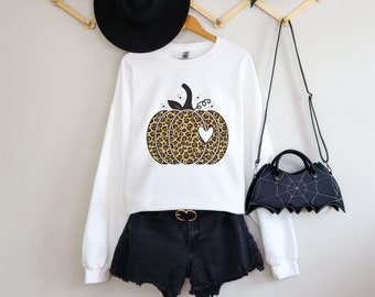 May include: A white sweatshirt with a leopard print pumpkin design, a black hat, and black shorts. A black bat-shaped purse hangs on the right. The pumpkin has a heart-shaped cutout.
