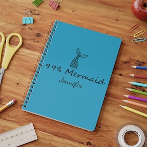 Può includere: Un quaderno a spirale turchese con un'illustrazione di coda di sirena nera e il testo "99% Mermaid Jennifer." Il quaderno è circondato da articoli di cancelleria, tra cui matite, forbici e graffette, su una scrivania di legno.
