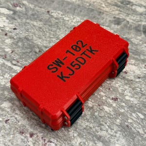 May include: A bright red rectangular case with the text "SW-102 KJ5DTK" printed in black. The case has black hinges and clasps. The case is closed and appears to be made of a durable material.