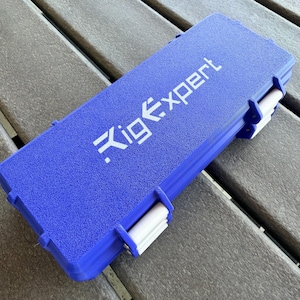 Puede incluir: Un estuche de transporte rígido azul de forma rectangular con una superficie texturizada y cierres blancos en el lateral. La palabra RigExpert está impresa en letras blancas en la superficie superior del estuche para su protección.