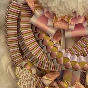May include: A decorative homecoming mum featuring layers of pleated pink, white, and gold glitter ribbons. It includes a white floral accent at the top and two cutouts with the text "five six seven eight" and "Class" in gold glitter script.