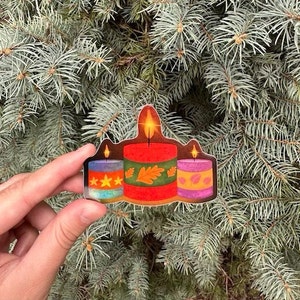 May include: Three candles with a fall theme. The candles are decorated with leaves and stars. The candle in the center is red, the candle on the left is blue and the candle on the right is purple.