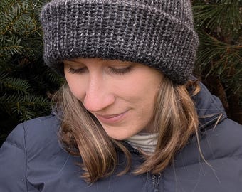May include: A gray, ribbed knit beanie hat. The hat is a dark gray color with a folded brim. The beanie is made of a soft, textured yarn. The hat is designed to keep the head warm in cold weather.