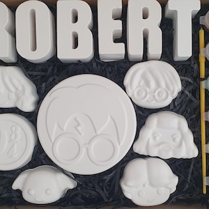 May include: A craft kit featuring the name "ROBERT" in white block letters. The kit contains white plaster figures of character faces and a round emblem, plus a set of paints and a brush for decorating.
