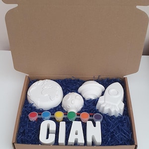 May include: A craft kit in a cardboard box, containing white space-themed figurines: a rocket, planet, moon, and another planet. The kit includes paint pots and the name "CIAN" in white block letters, set on blue shredded paper.