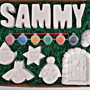 May include: A craft kit in a cardboard box, with the name "SAMMY" in large white letters. Includes paint pots, a paintbrush, and ceramic shapes such as a fairy door, sun, and ladybug. The box is filled with green paper.