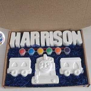 May include: A paint-your-own train set in a cardboard box. The set includes the name "HARRISON" in white block letters, a train engine, two train cars, paint pots, and paintbrushes. The box is filled with blue shredded paper.