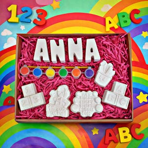 May include: A wooden box filled with pink shredded paper, containing white foam letters spelling "ANNA", paint pots, a paintbrush, and various foam shapes. The background features a rainbow and the numbers "123" and letters "ABC".