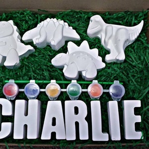 May include: A craft kit featuring four white dinosaur figurines, a set of paint pots, and the name "CHARLIE" spelled out in white letters. The dinosaurs and letters are ready to be painted.