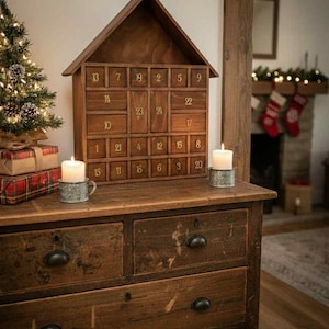 May include: A wooden advent calendar shaped like a house with numbered drawers. The calendar sits on a dark wood dresser with black hardware. Two candles in metal holders flank the calendar. A small Christmas tree and wrapped gifts are on the left.