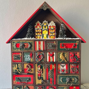 Puede incluir: Un calendario de adviento de madera con forma de casa y techo rojo. Cuenta con veinticuatro cajones individuales, cada uno decorado con motivos festivos únicos, herrajes metálicos y números. Edificios en miniatura y dos pinos coronan la estructura.