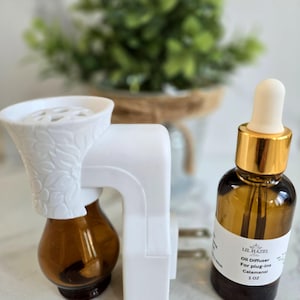 May include: White electric essential oil diffuser with a brown glass bottle and a white ceramic top.  A separate brown glass bottle with a gold cap and a white label that reads "Lil Hazel Oil Diffuser for plug-ins Calamansi 1 oz".