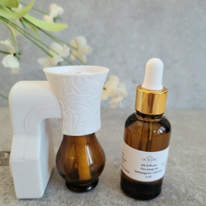 May include: White plug-in oil diffuser with a brown glass bottle and a white ceramic top. Next to it is a brown glass bottle with a gold lid and a white dropper. The bottle label reads "Lil Hazel Oil Diffuser For plug-ins Sampaguita - Jasmine 1 oz."