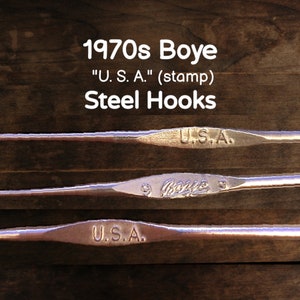 Könnte beinhalten: Drei vintage Boye Stahl-Häkelnadeln mit dem Text "U.S.A." auf zwei der Nadeln. Die Nadeln liegen auf einer Holzoberfläche.