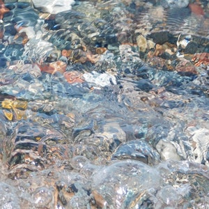 Könnte beinhalten: Nahaufnahme eines klaren Bachbettes mit glatten, bunten Steinen und Kieselsteinen, die unter dem welligen Wasser sichtbar sind.