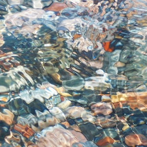 May include: A close-up of a stream bed with clear water flowing over smooth, colorful stones. The water creates a rippling effect, reflecting the light and colors of the stones.