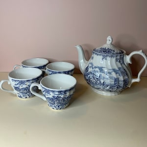 May include: A blue and white teapot and four teacups with a floral pattern. The teapot has a handle and spout. The teacups have handles. The teapot and teacups are on a white surface.
