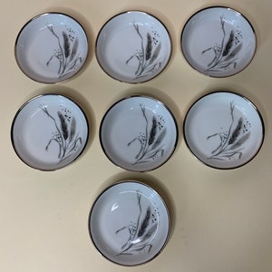 May include: Set of seven white ceramic saucers with a gold rim and a black and white wheat design.