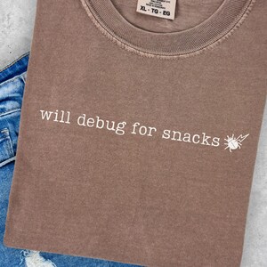Peut inclure: T-shirt marron Comfort Colors avec l'inscription "will debug for snacks" et un petit dessin d'insecte. Le t-shirt est plié sur un short en jean bleu.