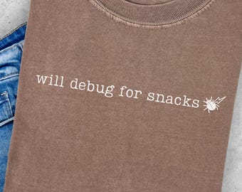 May include: A brown Comfort Colors t-shirt with the text "will debug for snacks" and a small graphic of a bug. The shirt is folded on top of a pair of blue denim shorts.