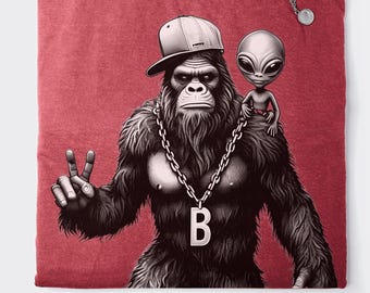 May include: A red t-shirt with a black and white graphic print of a gorilla wearing a baseball cap and a large chain with a letter 'B' pendant. A small grey alien is sitting on the gorilla's shoulder.
