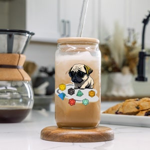 May include: A clear glass jar with a brown lid and a straw. The jar is filled with a brown liquid and has a sticker on the front featuring a cartoon pug wearing sunglasses and surrounded by colorful dice.