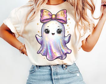 Puede incluir: Camiseta color crema con un simpático fantasma de dibujos animados con un gran lazo. El fantasma es blanco con un cuerpo morado y azul brillante, y el lazo es rosa y dorado. Estrellas doradas rodean al fantasma.