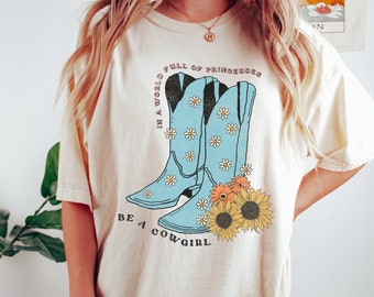 May include: A beige t-shirt with a graphic of blue cowboy boots with white daisies and the text "In a world full of princesses be a cowgirl".