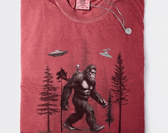 May include: Red t-shirt with a graphic print of Bigfoot carrying a small alien. The graphic also includes trees and flying saucers. The shirt has a Comfort Colors tag with the size XL, TG, and 60.