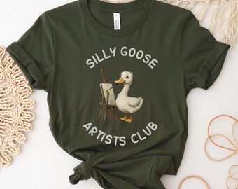 May include: A green t-shirt with the text "Silly Goose Artists Club" in white. A cartoon goose is holding a paintbrush and standing in front of an easel.