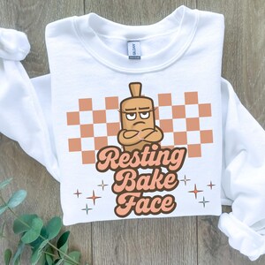 May include: White crew neck sweatshirt with a graphic design. The design features a cartoon rolling pin with crossed arms and the text "Resting Bake Face" in a retro font. A checkered pattern and star accents complete the design.