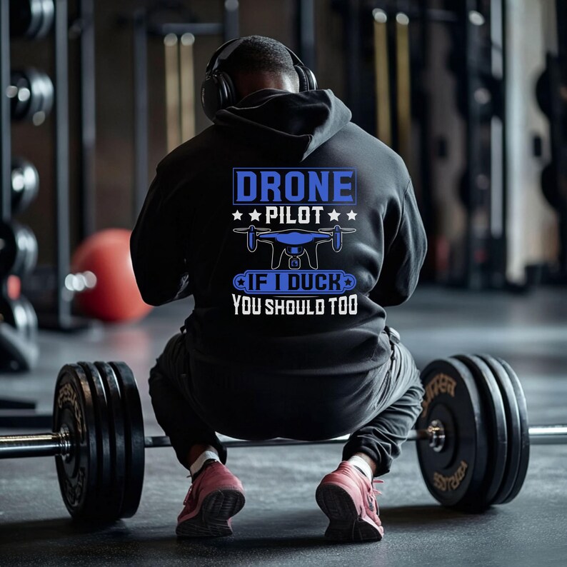 May include: A black hoodie with the text "DRONE PILOT" in blue and white, along with a drone graphic. The phrase "IF I DUCK YOU SHOULD TOO" is below the graphic. The person is wearing headphones and red sneakers, sitting in a gym.