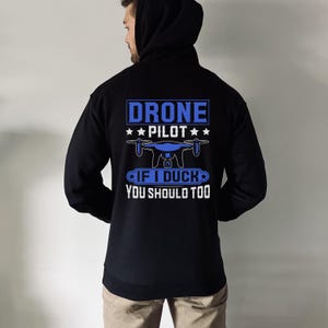 May include: Black hooded sweatshirt with blue text and a drone graphic. The text reads "DRONE PILOT" above a drone illustration, and "IF I DUCK YOU SHOULD TOO" below. The sweatshirt is worn by a person.