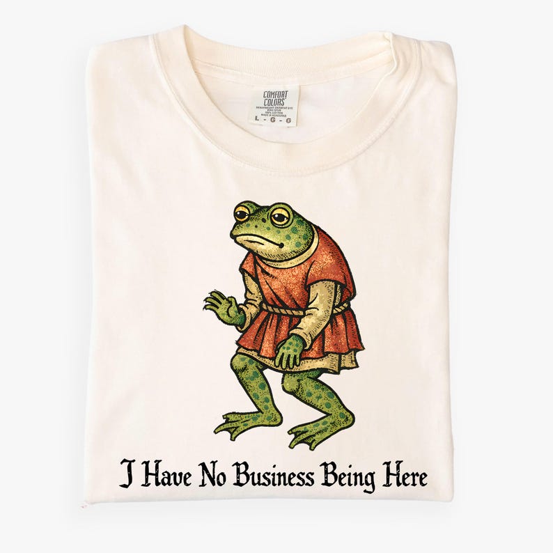 A t-shirt design showing a green frog standing upright in a medieval-style red tunic. Below the illustration is the phrase “I Have No Business Being Here” in black gothic-style lettering. The artwork has a vintage, illustrated appearance and is printed on a t-shirt.