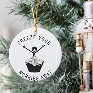 Könnte beinhalten: Weißer Keramik-Weihnachtsbaumschmuck mit einer schwarzen Strichmännchenfigur, die in einem Eimer mit Eiswürfeln steht. Der Schmuck trägt die Aufschrift "Freeze your worries away".