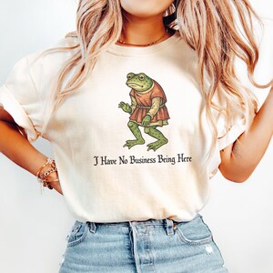 A t-shirt design showing a green frog standing upright in a medieval-style red tunic. Below the illustration is the phrase “I Have No Business Being Here” in black gothic-style lettering. The artwork has a vintage, illustrated appearance and is printed on a t-shirt.