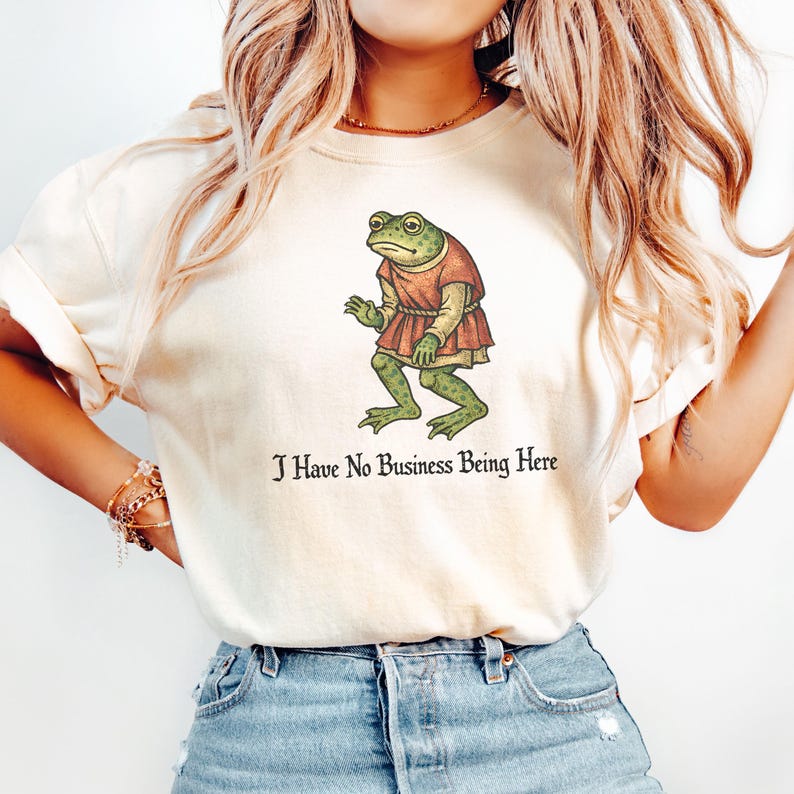 A t-shirt design showing a green frog standing upright in a medieval-style red tunic. Below the illustration is the phrase “I Have No Business Being Here” in black gothic-style lettering. The artwork has a vintage, illustrated appearance and is printed on a t-shirt.