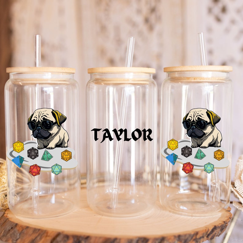 May include: Three clear glass tumblers with bamboo lids and straws. Each tumbler features a different design: one with a pug wearing sunglasses and surrounded by dice, one with the name "TAYLOR" in black text, and one with a pug wearing sunglasses and surrounded by dice.