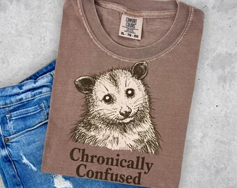 May include: A brown crew neck t-shirt featuring a black line art illustration of an opossum face. Below the opossum, the text reads "Chronically Confused" in a dark brown serif font. The shirt has a size XL tag visible at the neck.