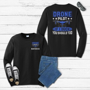 May include: Black long-sleeve shirts with the text "DRONE PILOT" and a drone graphic. One shirt has "MAVERICK" printed below the text. The back of another shirt has the text "IF I DUCK YOU SHOULD TOO". Includes blue jeans and black trainers.