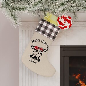 May include: A beige Christmas stocking with a black and white buffalo check cuff. The stocking features a cartoon cow with a red bow and the text "Mooney Christmas" and "Chelsie". A red and white swirled lollipop and a yellow gift are inside.