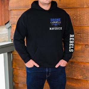 May include: Black hoodie with a front pocket and a hood. The front features the text "DRONE PILOT" above a drone graphic and the word "MAVERICK". The right sleeve has the word "REBELS" printed vertically in white.